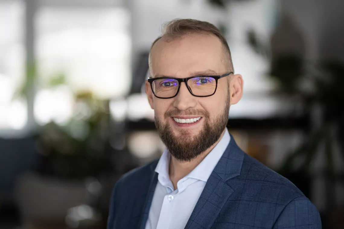 Mężczyzna w okularach, z krótkimi włosami i brodą, uśmiechnięty, ubrany w granatowy garnitur i jasnoniebieską koszulę. Tło jest rozmyte.