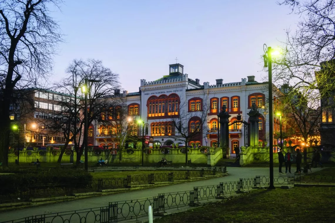 Zabytkowy budynek w otoczeniu zieleni, o zmroku, świecą się uliczne latarnie i okna w budynku