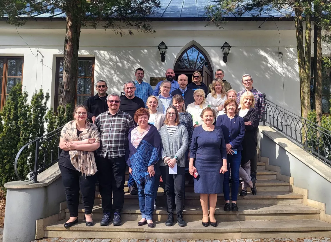 Grupa kilkunastu osób stojących na schodach przed białym budynkiem z łukowatymi oknami i drzwiami, wokół którego rosną drzewa. Osoby ustawione sa w kilku rzędach na schodach przed wejściem do budynku.