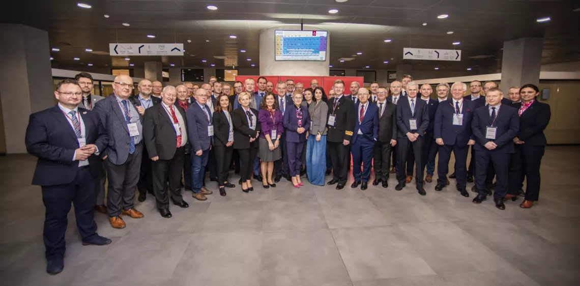 Duża grupa osób ubranych w formalne stroje, stojących razem w holu budynku, gdzie odbywa się konferencja