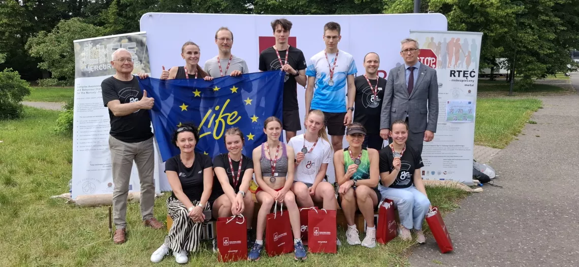 Grupa osób - część w sportowych strojach i z medalami na szyi stoi na tle banerów, dalej widac drzewa. Kilka osób trzyma unijną flagę z napisem life