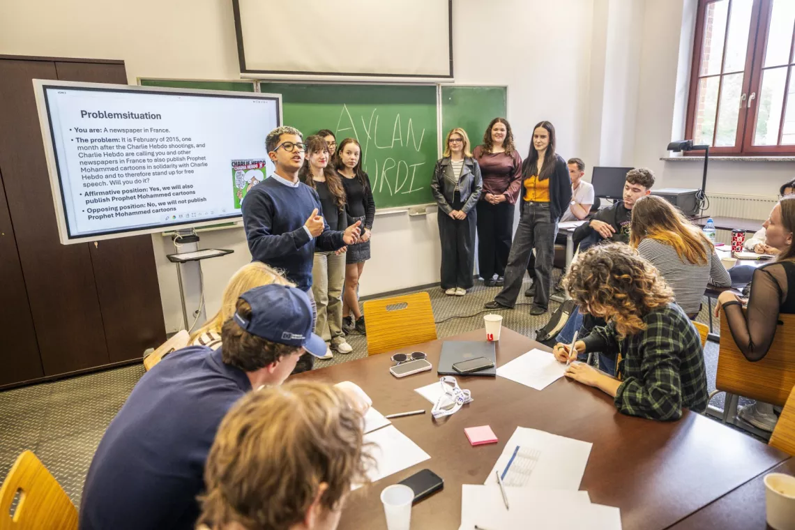 Debata prowadzona w ramach programu przrez moderatora stojącego przy ekranie. Wokół grupa studentów przy tablicy i przy stołach