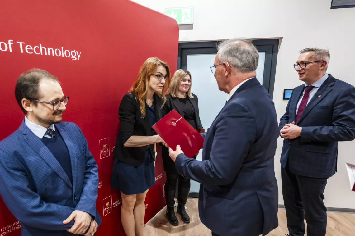 Dwaj elegancko ubrani mężczyżni wręczają i składają gratulacje kobiecie w okularach i z długimi włosami, która stoi w szeregu z dwiema innymi osobami: kobietą i mężczyzną
