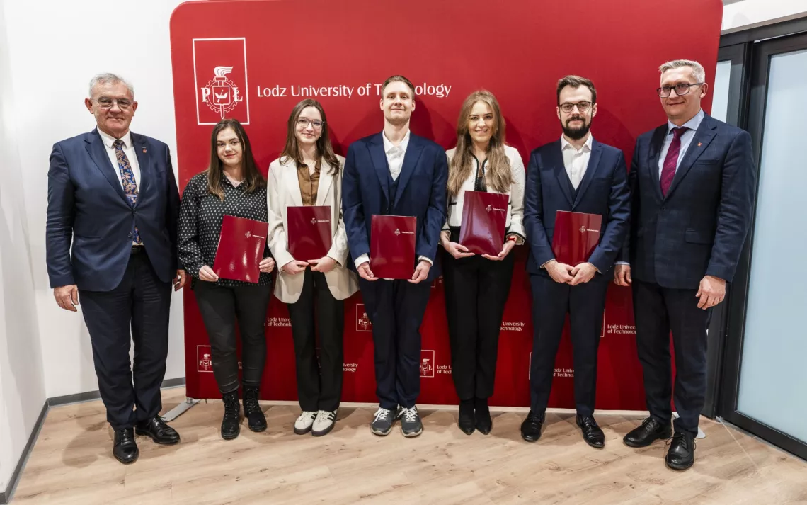 Laureatki i laureaci stypendium profesorskiego trzymają w rękach teczki na dokumenty, obok stoją dwaj mężczyni w garniturach