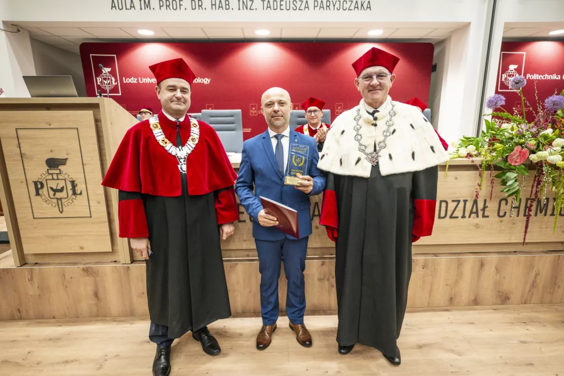 Mężczyzna w eleganckim garniturze trzymający statuetkę w ręku stoi między prorektorem i rektorem