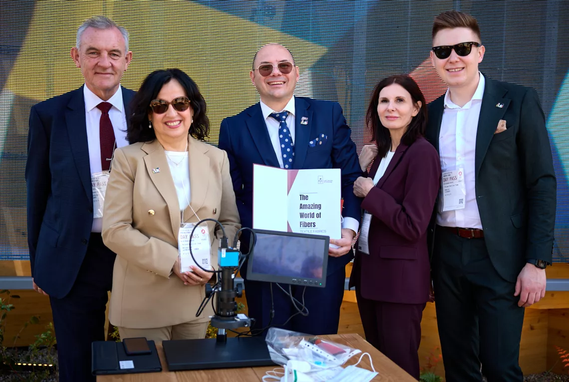 Pięć osób stoi za stołem, na którym znajdują się różne przedmioty: mikroskop, laptop i notatki