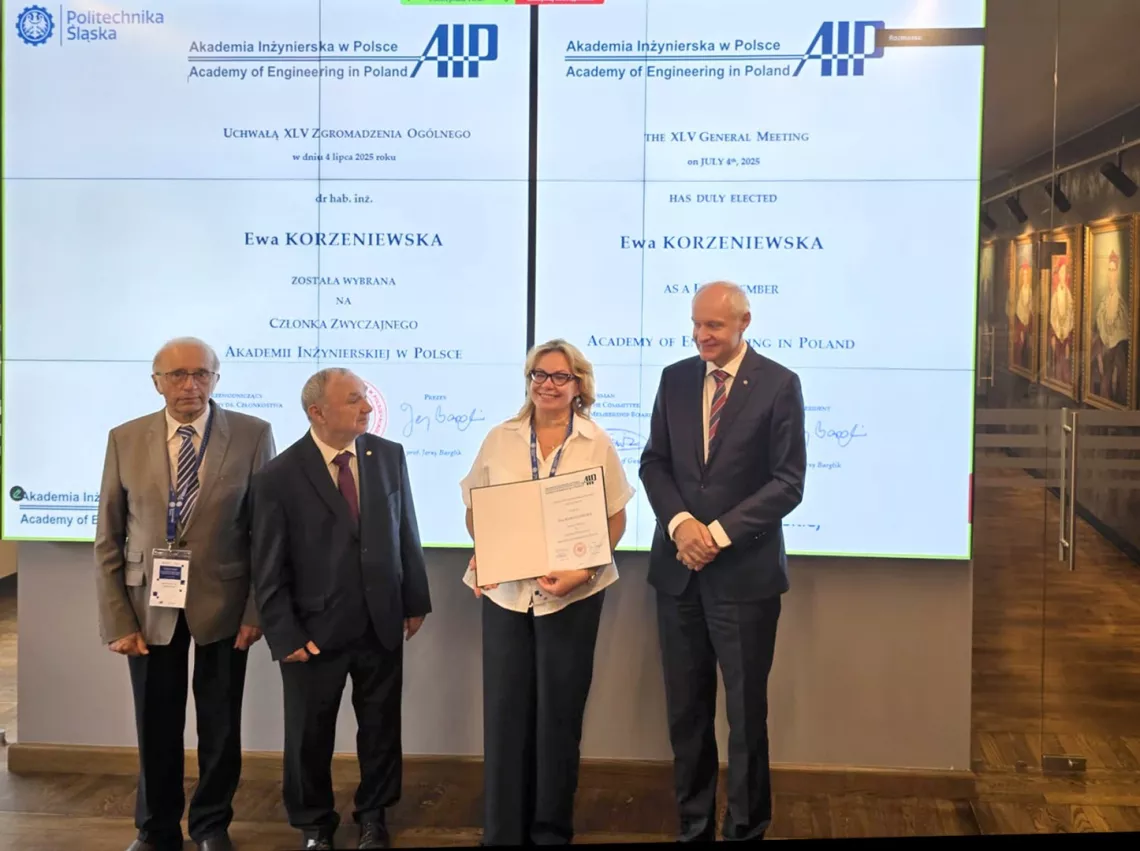 Kobieta z dyplomem w ręku stoi w towarzystwie trzech mężczyzn. Z tyłu ekran, na którym wyświetla się treść dyplomu.