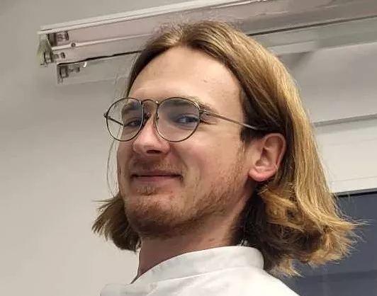 Twarz młodego mężczyzny w okularach i z dłuższymi włosami zaczesanymi za uszy.