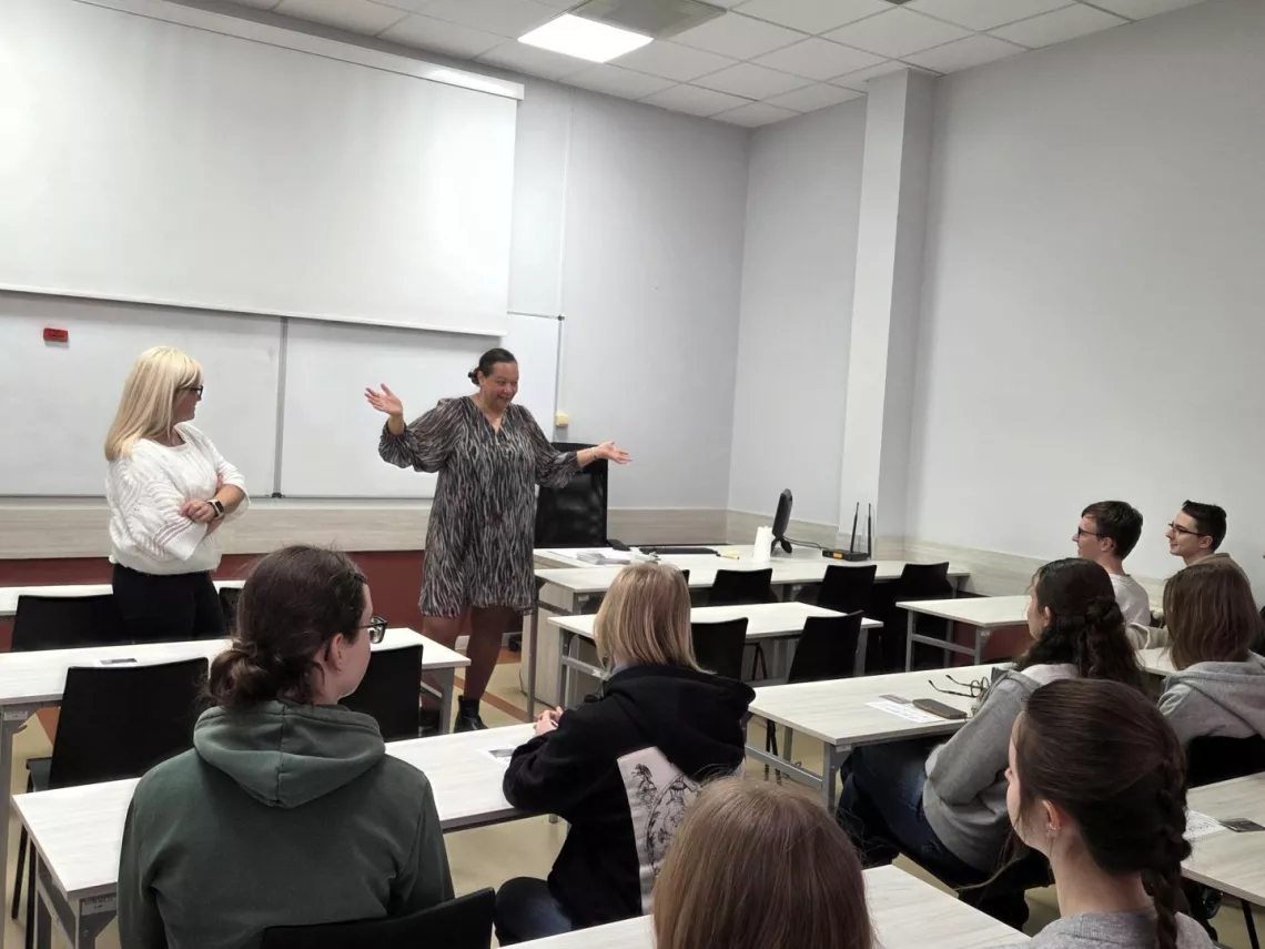 Studenci siedzą w autytorium przy stolikach, słuchając wykładu kobiety