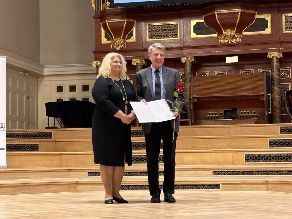 Mężczyzna trzymający dyplom i różę stoi obok kobiety na scenie reprezentacyjnej auli