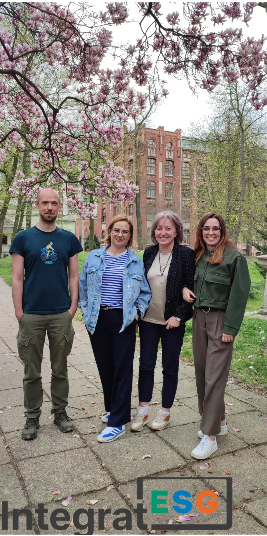 Trzy kobiety i mężczyzna stoją przy kwtnącym drzewie, z tyłu widać budynek z czerwonej cegły. Pod spodem jest napis Integrat ESG