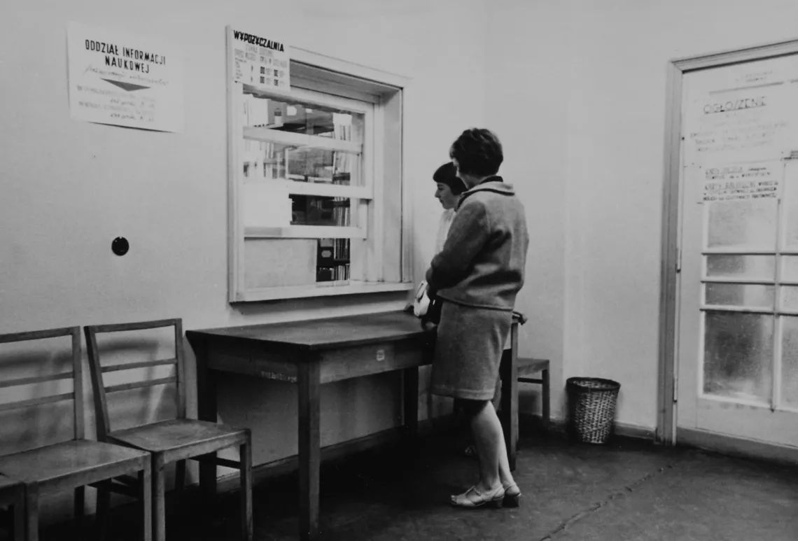 Dwie kobiety przy okienku wypożyczalni książek w Bibliotece Politechniki Łódzkiej, rok 1962. Wnętrze z prostymi meblami i tablicami informacyjnymi.