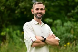 Mężczyzna z brodą w jasnej koszuli trzyma ręce skrzyżowane na piersiach, wokół dużo zieleni