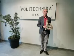 Mężczyzna w okularach, szarej marynarce i kolorowej koszuli trzyma w rękach odlee, stojąc na tle ściany, fo której przymocowana jest tablica z napisem Politechnika 1945odle 