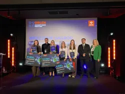Laureaci konkursu podczas uroczystości wręczenia nagród. Trzymają w rękach symboliczne czeki, w tle ekran logo Orlenu, z boku oświetlenie sceny
