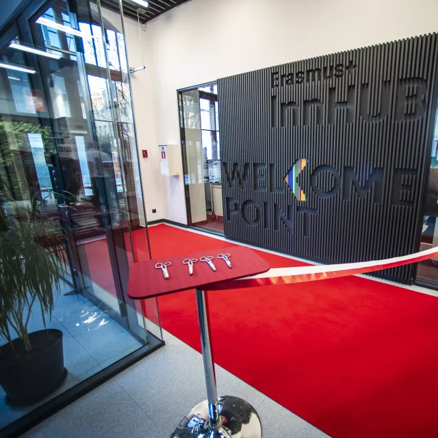 Przestzreń z duzym napisem Erasmus+ InnHub umieszconym na ścianie, rozłożonym czerwonym dywanem, wstęgą do przecięcia.