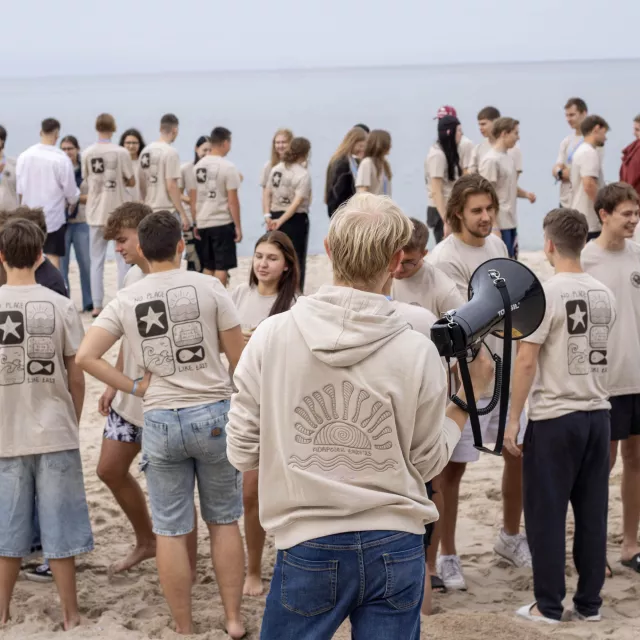 Grupa młodych osob ubranych w jednakowe podkoszulki jest na plazy, rozmawia, a na pierwszym planie stoi osoba z megafonem