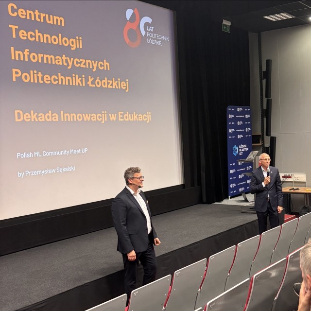 Dwóch mężczyzn stoi przed publicznością siedzącą na widowni, jeden z nich przemawia do mikrofonu