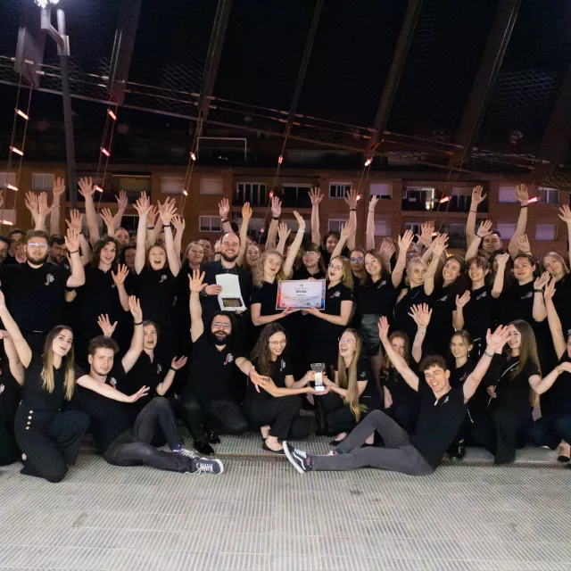 Grupa ludzi ubranych na czarno pozuje do wspólnego zdjęcia, unosząc ręce w geście radości. W tle widoczne są budynki oraz dwa banery reklamowe.