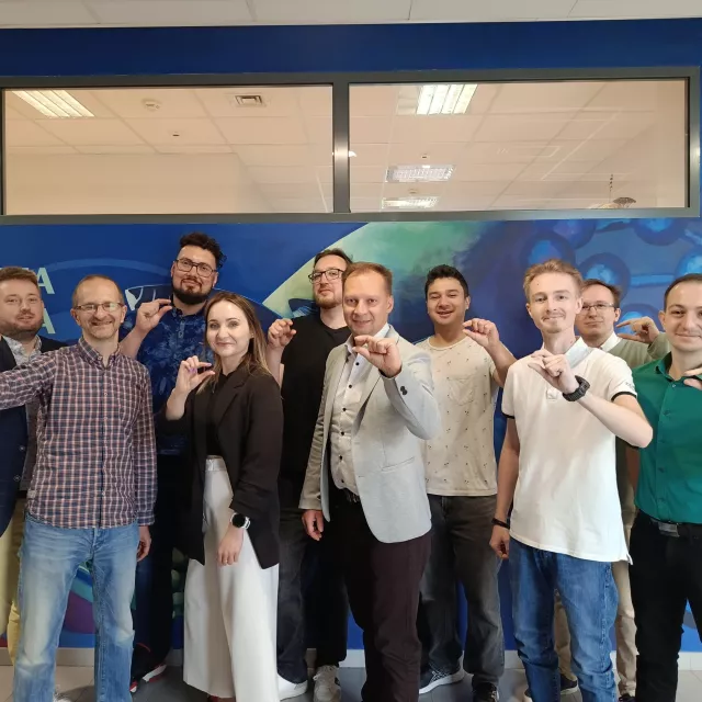 Grupa osób wykonuje gest palcami pokazujący, że coś jest bardzo małe. Osoby stoją na tle ściany z napisem inżynieria materiałowa.