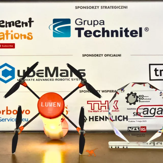 Świecący dron, obok stoi nagroda w formie szklanej plakiety, z tyłu plansza z nazwami sponsorów i ich logo