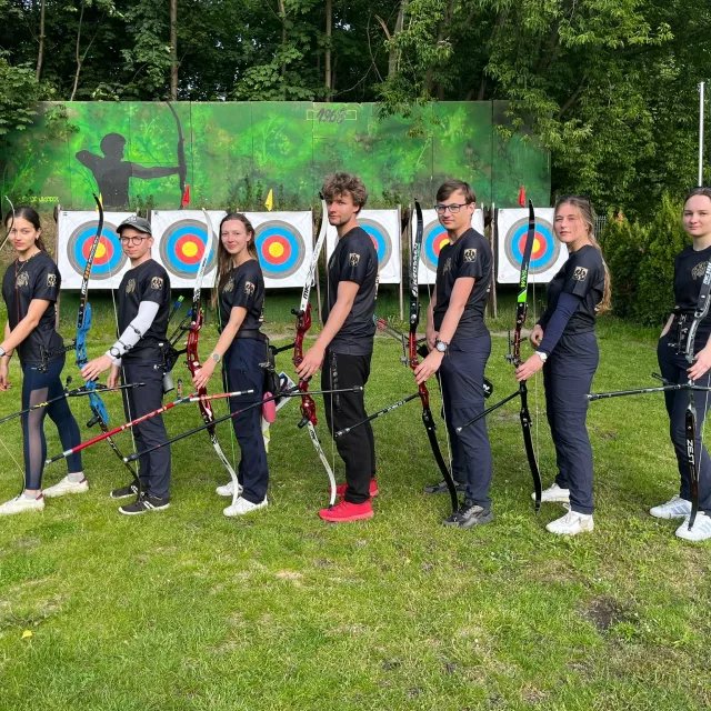 Siedmioro zawodników stoi z łukiem w ręku, w tle tarcze strzelnicze