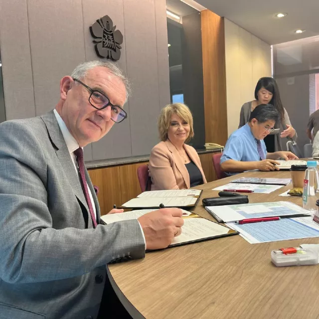 Elegancko ubrany mężczyzna podpisuje dokument, przy stole siedzi jeszcze kilka osób.