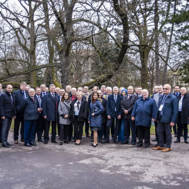 Grupa osób stoi na zewnątrz w parku. W tle drzewo bez liści.