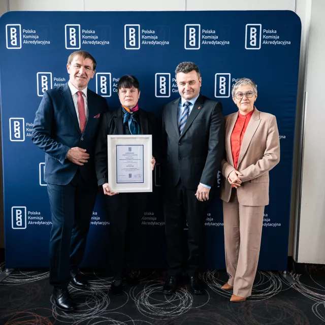 Dwie kobiety - jedna z nich trzyma dyplom - i dwóch mężczyzn stoi na tle ścianki z napisem Polska Komisja Akredytacyjna