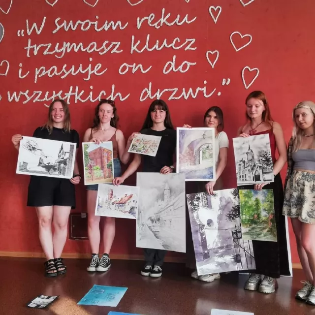 sześć młodych kobiet z pracami stoi i trzyma plastyczne prace w reku. Z tyłu napis na czerwonym tle: W swoim ręku trzymasz klucz i pasuje on do wszystkich drzwi.